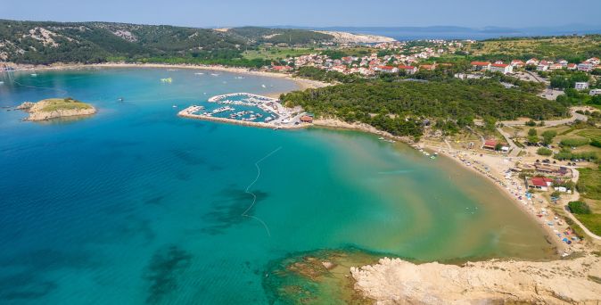 Luftaufnahme des Rajska Plaža Strands auf der Insel Rab in Kroatien mit langer Sandbucht, türkisblauem Meer, Yachthafen und Küstenort im Hintergrund