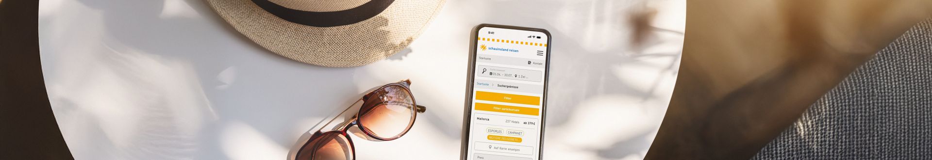 Smartphone mit geöffneter Reisebuchungsseite neben Sonnenbrille und Strohhut auf hellem Tisch