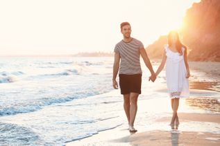 Paar geht Hand in Hand am Strand entlang, während sanfte Wellen ans Ufer rollen und warmes Abendlicht die Szene in goldenem Glanz einfärbt