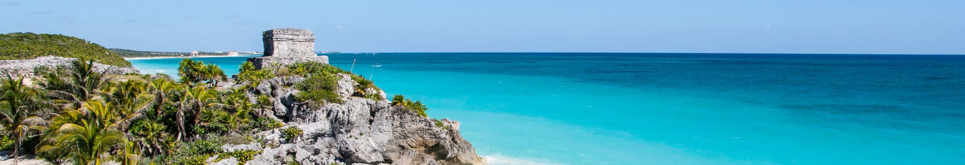 Blick auf die Küste von Mexiko mit türkisfarbenem Wasser, Felsen und Palmen im Vordergrund. Im Hintergrund ist ein historischer Turm sichtbar.