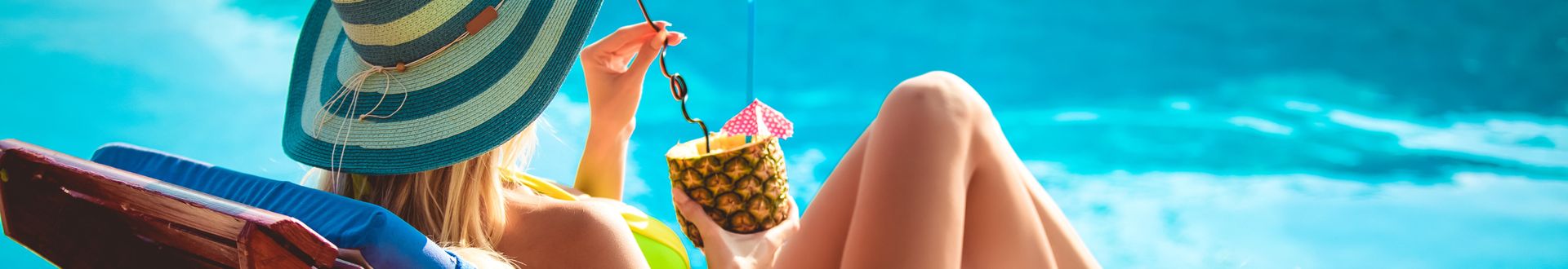 Frau mit gestreiftem Sonnenhut und gelbem Bikini entspannt auf Sonnenliege am Pool und hält Ananas-Getränk mit Strohhalm und kleinem Schirmchen