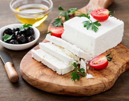 Frischer Feta-Käse in Scheiben auf rustikalem Holzbrett mit Oliven, Tomaten, Petersilie und Olivenöl