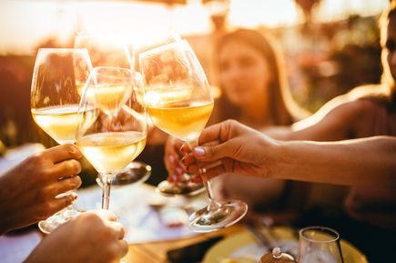 Mehrere Personen stoßen mit Gläsern Weißwein bei Sonnenuntergang an, geselliges Beisammensein im Freien