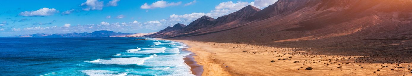 Strand mit Wellen und Vogel am Himmel, rechts Sanddünen und Berge bei Betancuria auf Fuerteventura unter blauem Himmel mit wenigen Wolken.