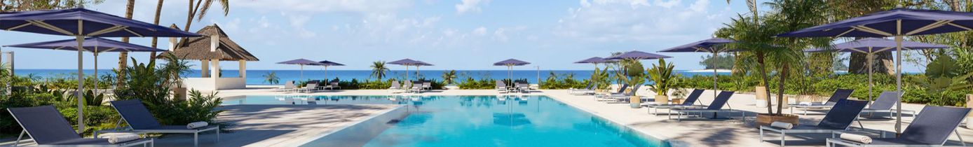 Großer rechteckiger Pool mit Liegestühlen und Sonnenschirmen an beiden Seiten, umgeben von Palmen und Blick auf das Meer im Hintergrund