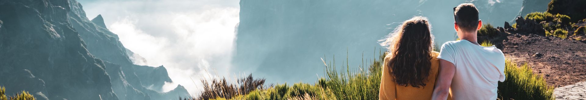Zwei Personen sitzen am Aussichtspunkt Pico do Arieiro auf Madeira und blicken über eine Berglandschaft mit steilen Felsformationen und Wolken zwischen den Gipfeln
