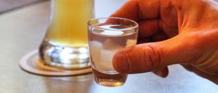 Hand hält ein kleines, klares Schnapsglas mit transparentem Inhalt, im Hintergrund unscharf ein Bierglas auf einem Untersetzer.