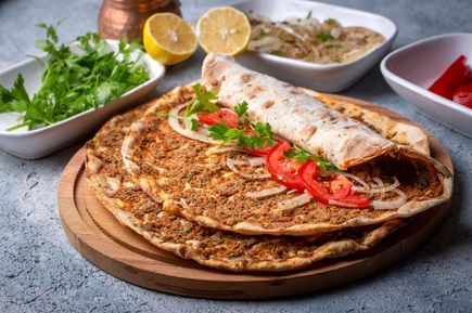 Lahmacun auf Holzbrett, belegt und gerollt mit Tomaten, Zwiebeln und Petersilie, daneben Zitronen und Kräuter