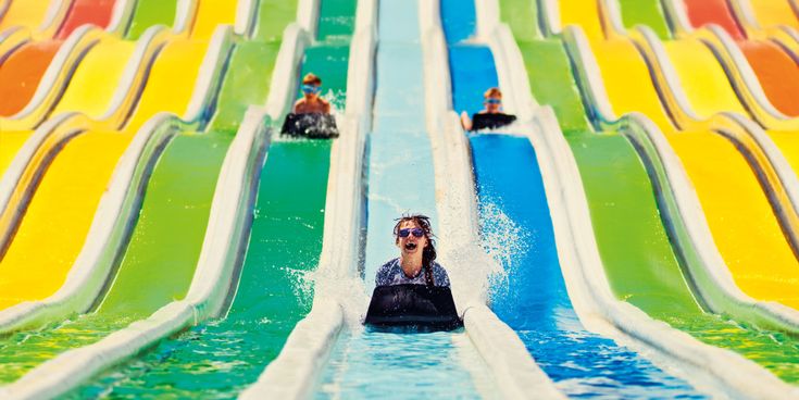 Drei Kinder rutschen auf Matten eine bunte Mehrfach-Wasserrutsche hinunter, Wasser spritzt, Spaß im Wasserpark