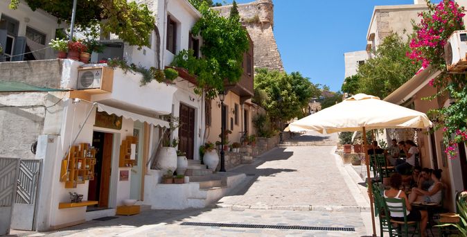 Schmale Gasse in Rethymnon auf Kreta mit weiß getünchten Häusern, blühenden Bougainvillea und Menschen unter einem Sonnenschirm an einem Straßencafé.