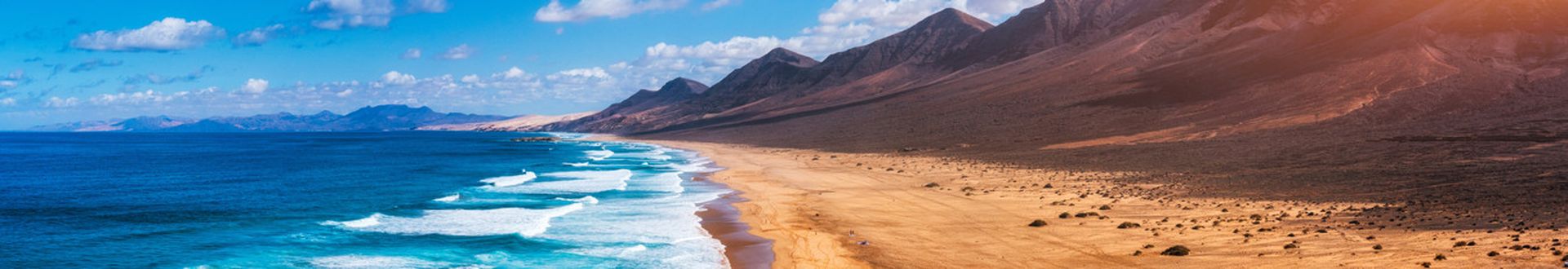 Strand mit Wellen und Bergen im Hintergrund unter blauem Himmel mit vereinzelten Wolken