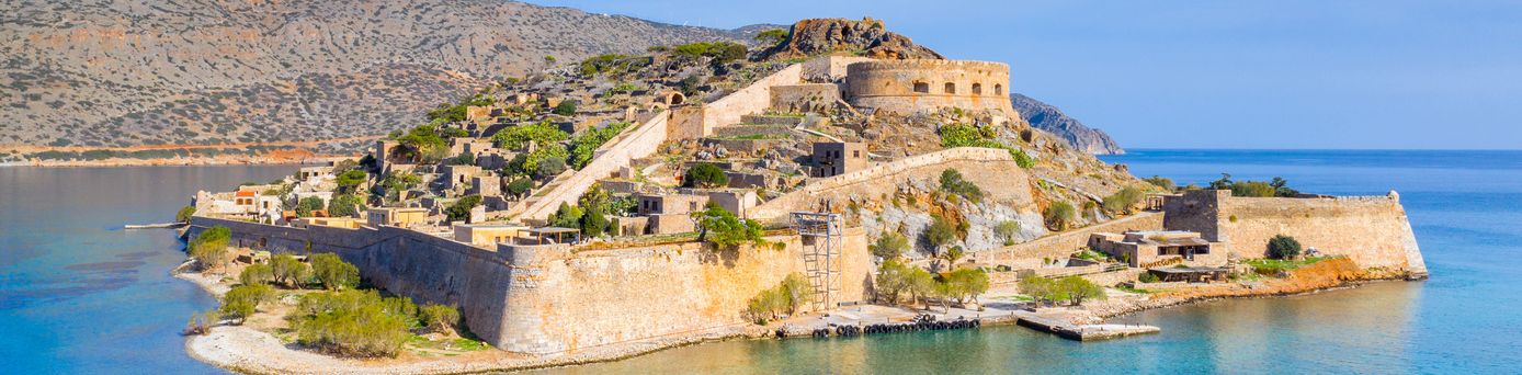 Blick auf eine historische Festung auf einer Halbinsel mit Mauern, Türmen und Gebäuden, umgeben von Wasser und karger Vegetation.