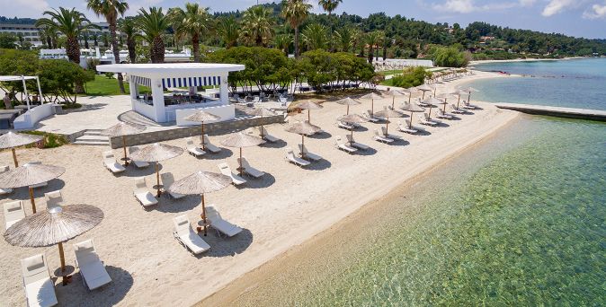 Sandstrand des Kassandra Palace Seaside Resort mit Liegen und Sonnenschirmen am klaren, flachen Meerwasser sowie einer Beachbar zwischen Palmen.