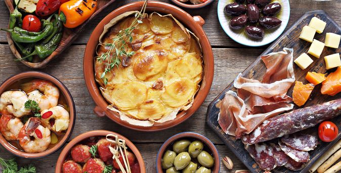 Verschiedene Tapas mit Kartoffelgratin, Oliven, Chorizo, Schinken, Käse, gegrilltem Gemüse und Garnelen auf Holztisch angerichtet