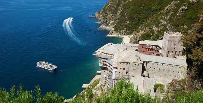 Blick von oben auf ein großes Klostergebäude an einer steilen Küste, davor ein Ausflugsboot auf blauem Wasser mit sichtbarer Bugwelle.