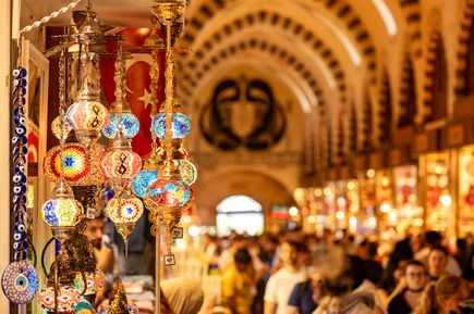 Detailaufnahme farbenfroher orientalischer Hängelampen im Gewürzbasar Istanbul, dahinter die belebte Markthalle mit warmem Licht und vielen Besuchern