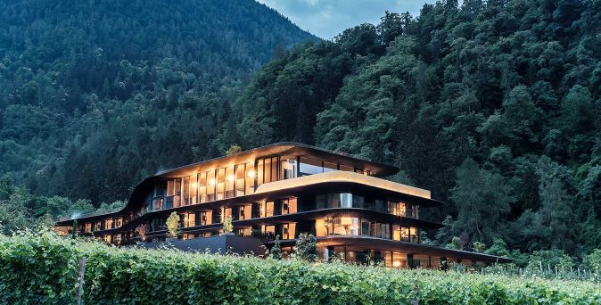 Modernes Smart Hotel Prinz Rudolf mit geschwungener Architektur, großen Glasfronten und warm beleuchteten Räumen, eingebettet in eine bewaldete Berglandschaft mit Weinbergen im Vordergrund.