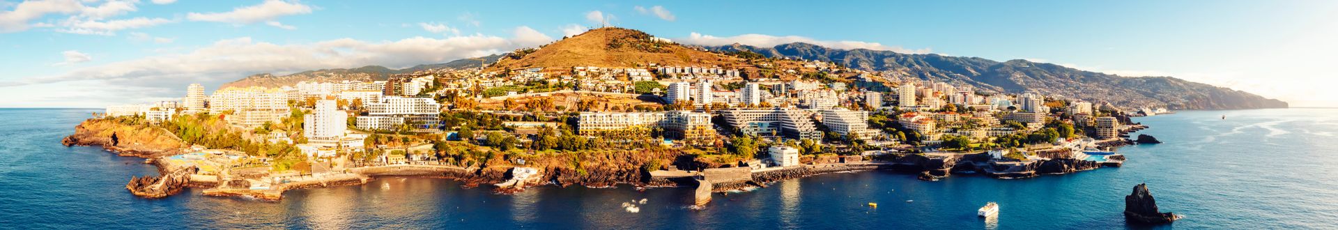 Panorama der Küste von Funchal auf Madeira mit dicht bebautem Hang, Hotels und Wohngebäuden entlang der felsigen Atlantikküste sowie Bergen im Hintergrund unter blauem Himmel