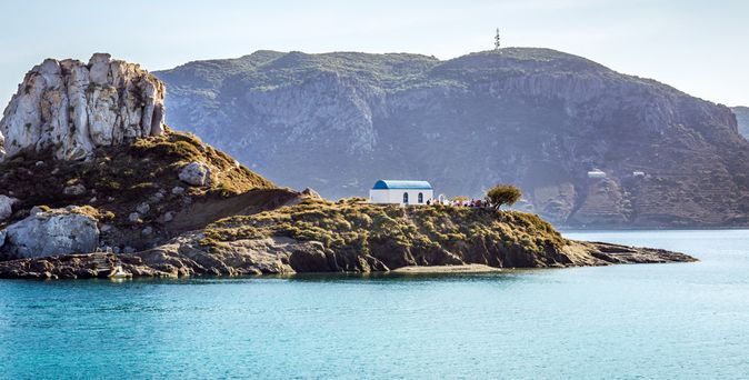 Kleine felsige Insel mit weiß-blauem Gebäude im Meer vor bewaldetem Berg im Hintergrund