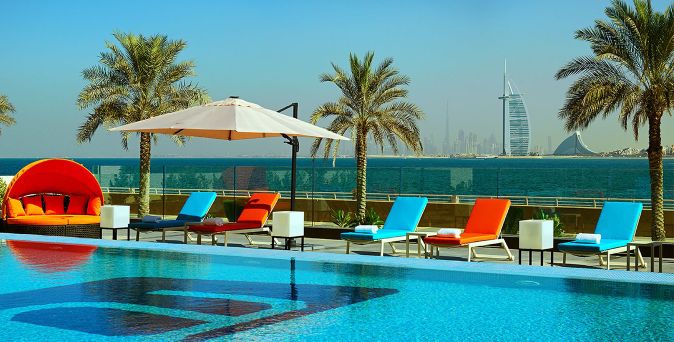 Poolbereich des Aloft Palm Jumeirah Hotels mit Liegestühlen in Blau und Orange, Sonnenschirm und Palmen, im Hintergrund das Meer und der Burj Al Arab in Dubai.