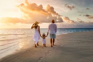 Familie mit einem kleinen Kind geht bei Sonnenuntergang am Strand entlang und schaut aufs Meer, während warme Abendwolken den Himmel färben