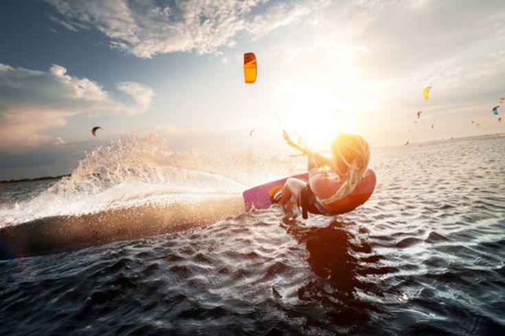 Kitesurfer gleitet dynamisch über das Wasser, während im Hintergrund mehrere Kites in der Luft schweben, Sonnenuntergangsstimmung
