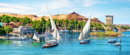 Drei traditionelle Feluken mit weißen Segeln segeln auf dem Nil vor grünen Ufern und sandigen Hügeln mit Gebäuden