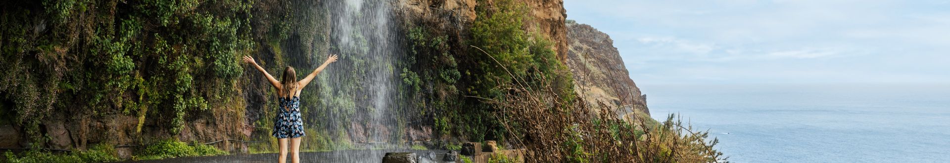 Person steht vor dem Wasserfall Cascata dos Anjos auf Madeira mit ausgebreiteten Armen zwischen einer bewachsenen Felswand und der Küste des Atlantiks