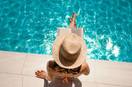 Frau mit Strohhut sitzt am Poolrand und liest ein Buch, während die Sonne auf das türkisblau glitzernde Wasser scheint