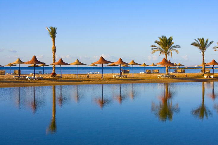 Strand in Hurghada am Roten Meer, Ägypten, mit Reihen von Sonnenschirmen, Liegen und Palmen, spiegelndes Wasser im Vordergrund unter klarem blauem Himmel