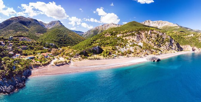 Bucht mit Sandstrand und klarem Wasser vor bewaldeten Hügeln und Bergen unter blauem Himmel mit Wolken