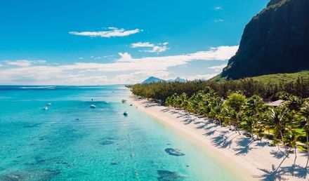 Langer Sandstrand auf Mauritius mit klarem türkisfarbenem Meer, flachen Wellen, Booten im Wasser und dichter grüner Vegetation vor einer Bergkulisse