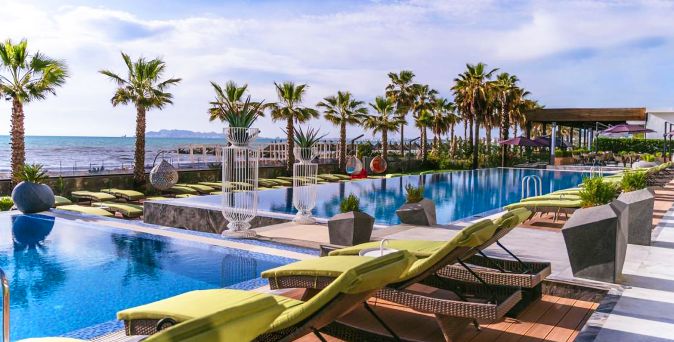Außenpool des Pinea Hotel Resort & Spa mit modernen Sonnenliegen, Palmen und direkter Lage am Meer vor der Küstenkulisse im Hintergrund