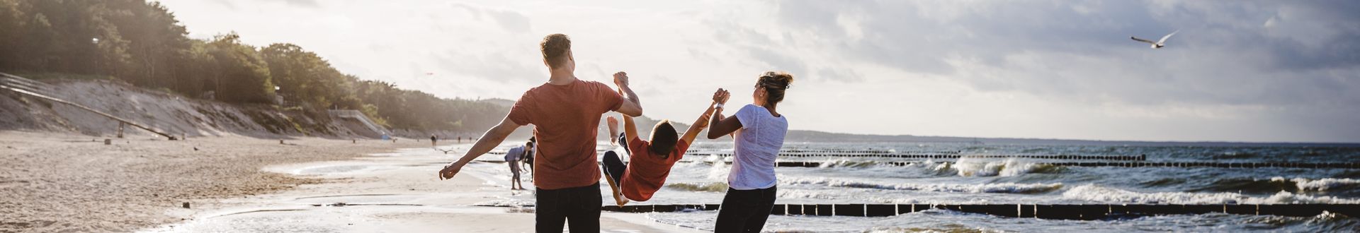 Zwei Erwachsene schwingen ein Kind am Strand mit Wellen und Dünen im Hintergrund