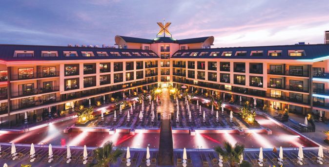 Das Hotel The X Belek mit beleuchtetem Poolbereich und mehreren Etagen mit Balkonen bei Dämmerung