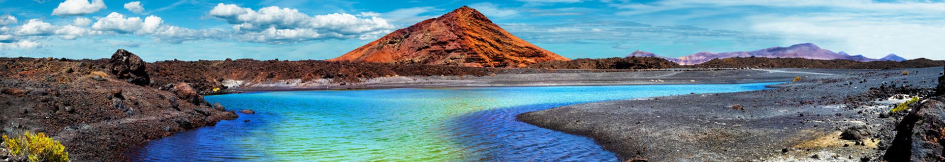 Berg mit rötlicher Spitze hinter einem See mit blau-grünem Wasser und Ufer mit schwarzem Lavagestein und gelblichen Pflanzen