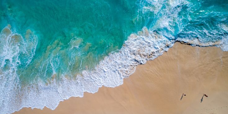 Luftaufnahme von Wellen, die an einen Sandstrand rollen, mit zwei Personen, die am Wasser entlanglaufen