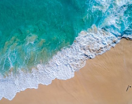 Luftaufnahme von Wellen, die an einen Sandstrand rollen, mit zwei Personen, die am Wasser entlanglaufen