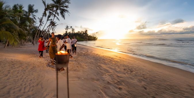Gruppe von Menschen am Strand bei Sonnenuntergang mit brennender Fackel im Vordergrund und Palmen im Hintergrund