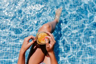 Frau sitzt entspannt am Poolrand und hält ein Getränk über dem klaren Wasser eines gefliesten Hotelpools