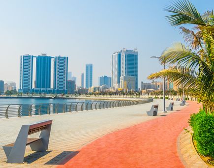 Promenade mit Bänken und Palmen am Wasser, im Hintergrund moderne Hochhäuser in Ajman
