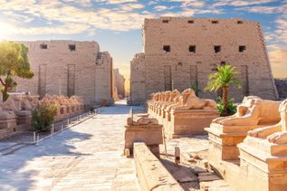 Blick auf den Karnak-Tempel in Ägypten mit Reihen von liegenden Sphinx-Statuen aus Stein und großen Sandsteinmauern im Hintergrund
