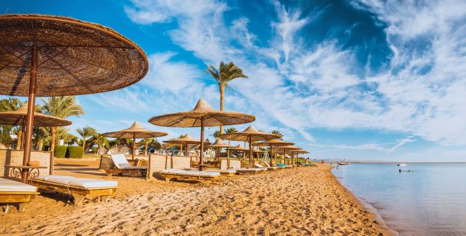 Sandstrand am Roten Meer in Ägypten mit Sonnenliegen, Schilfsonnenschirmen, Palmen und ruhigem, türkisfarbenem Wasser unter blauem Himmel