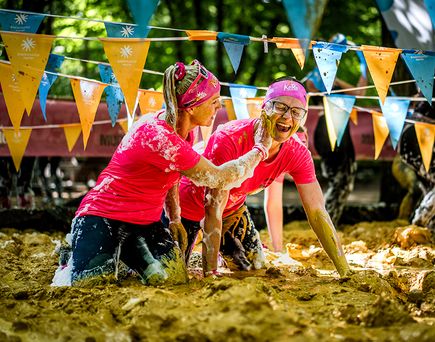 Teilnehmende des Muddy Angel Runs bewegen sich kriechend durch ein schlammiges Hindernis, während eine Person einer anderen Schlamm ins Gesicht streicht