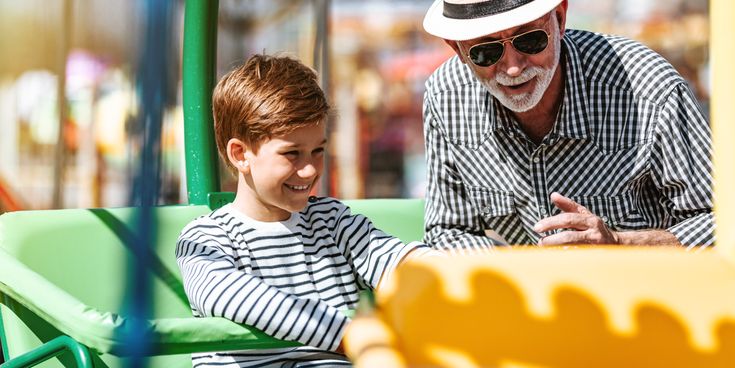 Junge sitzt lächelnd in einem Freizeitpark-Fahrgeschäft, begleitet von einem älteren Mann mit Hut und Sonnenbrille
