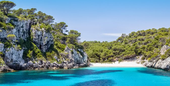 Klares türkisfarbenes Wasser in einer Bucht umgeben von steilen Felsen und grünen Kiefern auf Menorca