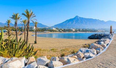 Ruhiger Sandstrand in Marbella, Spanien, mit Palmen, Felsenpromenade und Blick auf ein Bergpanorama am klaren Mittelmeer
