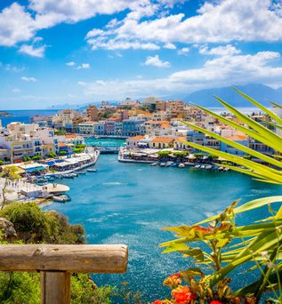 Blick auf den Hafen von Agios Nikolaos auf Kreta mit bunten Häusern, blauem Meer und lebendiger Uferpromenade unter sonnigem Himmel