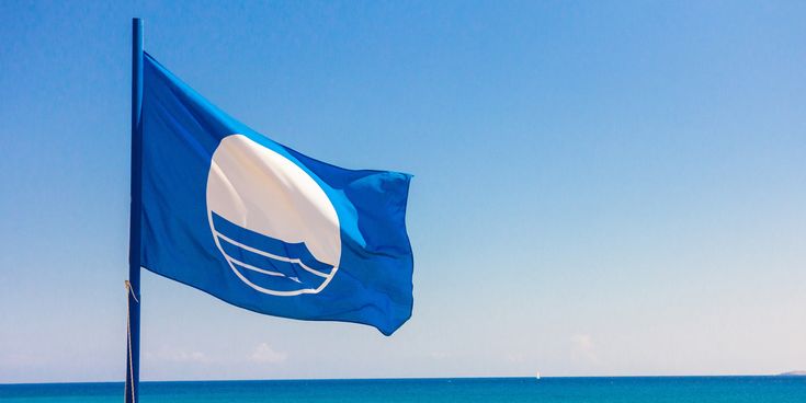 Blaue Flagge weht im Wind vor klarem Himmel über dem türkisblauen Meer als Symbol für ausgezeichnete Wasserqualität und Umweltstandards