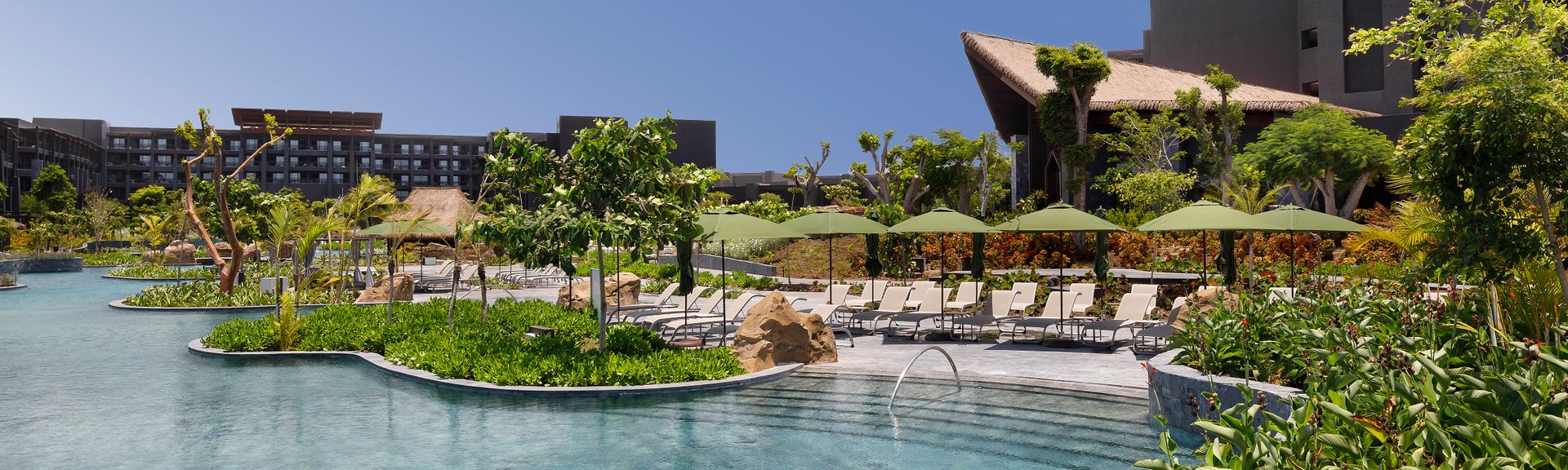 Poollandschaft mit Liegestühlen, Sonnenschirmen und begrünten Inseln vor mehrstöckigen Gebäuden